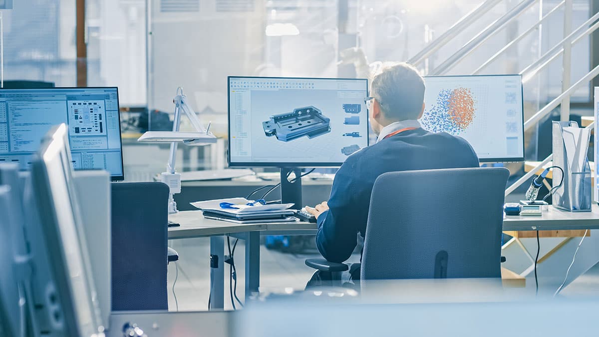 An engineering office, a designer modelling a machined part in CAD across two screens.