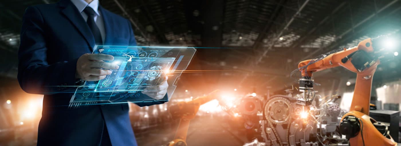 Production data on a handheld tablet, with robotic welding arms working an engine assembly on the line beyond.