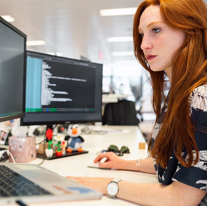 A project engineer working at a desk with a monitor and laptop in an open plan office.